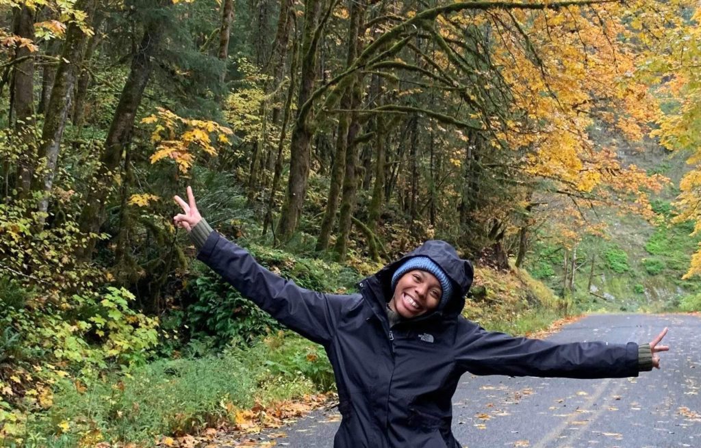 Lisa posing with arms out on an empty road behind thick forest with yellow, orange, and green leaves. 